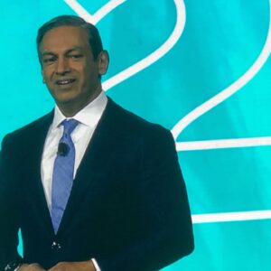 Sherrif Karamat speaking onstage at PCMA in front of a teal backdrop