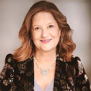 A portrait of Debra Zabloudil. She is a white woman with wavy brown hair and a black floral suit jacket