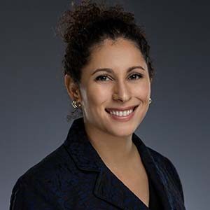 A portrait of Raquel Sharma. She is a brown woman with curly hair in a ponytail and a blue jacket