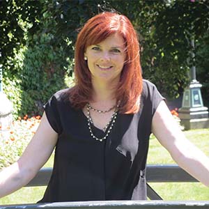 A portrait of Amie Devine. She is a white woman with straight red hair and a black short-sleeved blouse