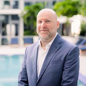 A portrait of Duncan Clements. He is a bald white man with a stubbled beard and a blue suit standing at a pool