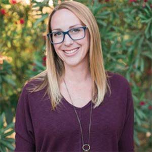 A portrait of Jamie Snively. She is a blonde white woman with square glasses and a dark purple blouse