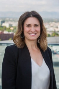 A portrait of Rachel Bendick. She is a white woman with wavy highlighted hair and a black suit jacket
