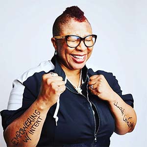 A portrait of Tess Vismale. She is a black woman with a mohawk, cat eye glasses and a dark blue jacket. She has "empowering with intent" written on one arm