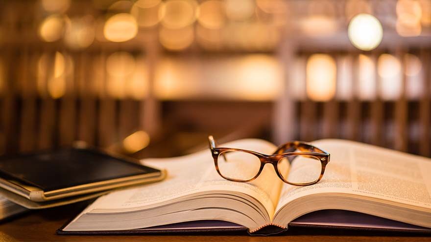 An open books with a pair of brown glasses on top. A shelf behind holds more books