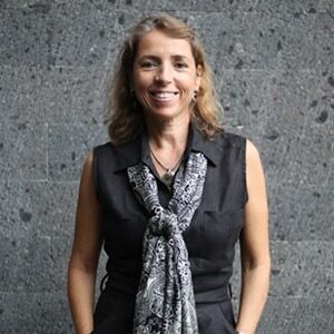 A portrait of Ana Henriques. She is a woman with blonde wavy hair and a sleeveless grey collared shirt