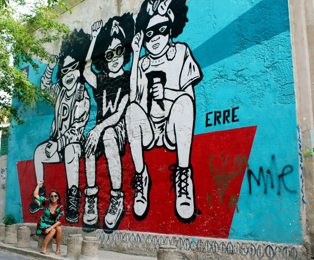 Ashley Lawson posing in front of a mural in Erre. The mural shows three black girls sitting on a red ledge with fists raised casually