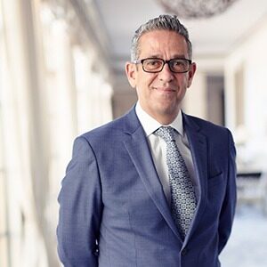 A portrait of Rodrigo Formoso. He is a brown man with combed greying hair, square glasses and a blue suit