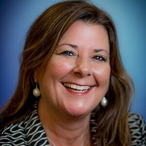 A portrait of Gina Gemberling Wibben. She is a white woman with wavy brown hair and a black patterned blouse