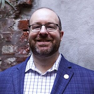 A portrait of Michael Klein. He is a balding white man with square glasses, a stubbled beard and a blue plaid suit