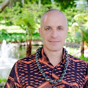 A portrait of Mike Kass. He is a balding white man with a dark Hawaiian shirt and a black nut lei