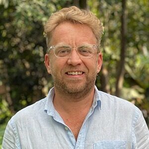 A portrait of James Wyndham. He is a blond white man with clear glasses, a stubbled beard and blue collared shirt