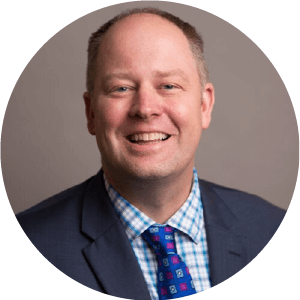 A portrait of Dr. Eric Olson. He is a balding white man with a dark suit and a blue patterned tie