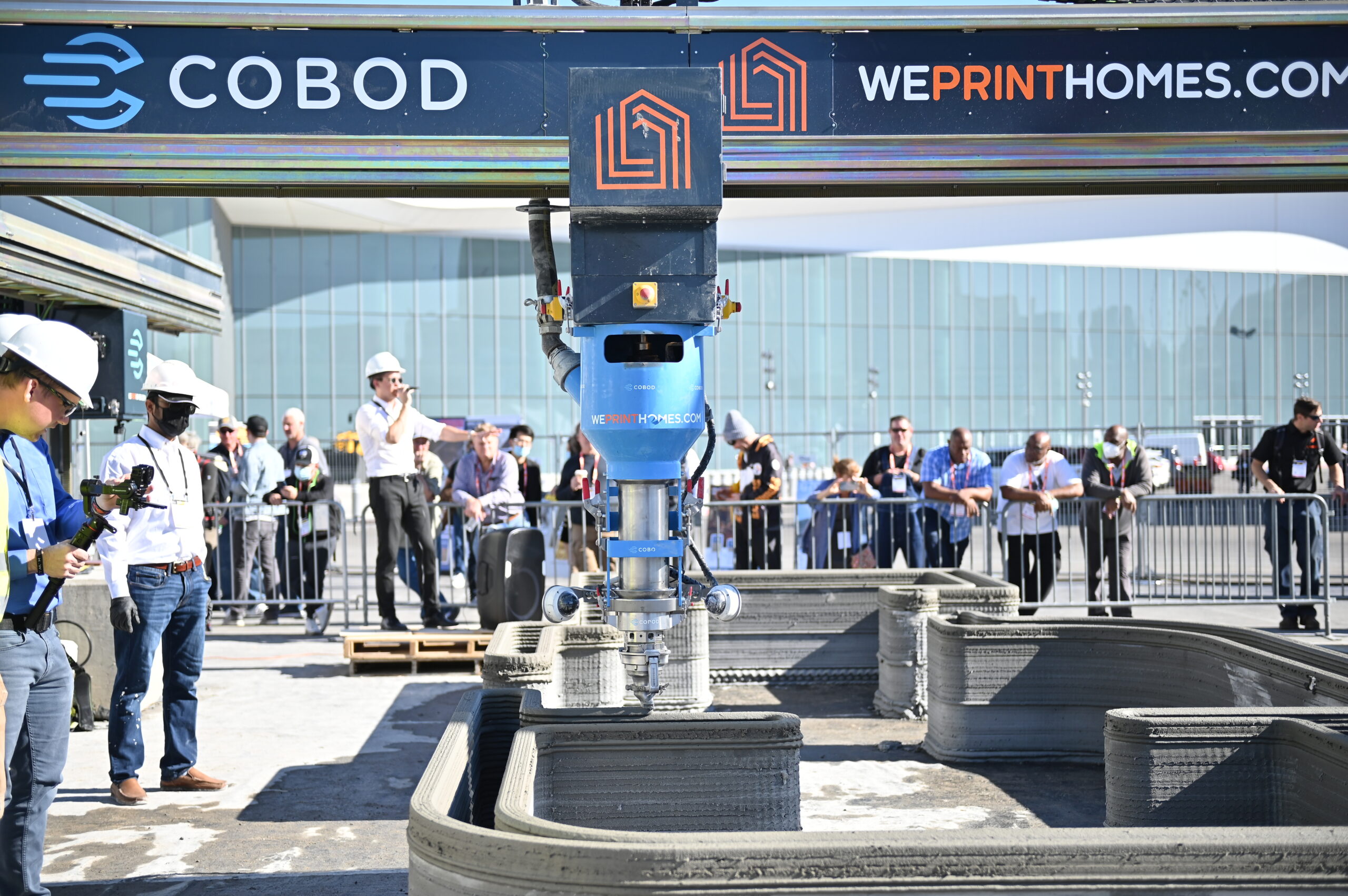 A demonstration of a large 3D printer making a house. Text on the arm of the printer reads "Cobod" and "We print homes.com"