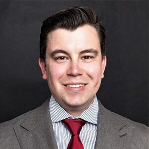 A portrait of Brock Taylor. He is a white man with combed black hair and a grey suit.