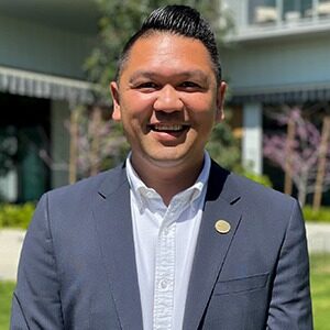 A portrait of Matthew De Guzman. He is a South Asian man with combed black hair and a grey suit.