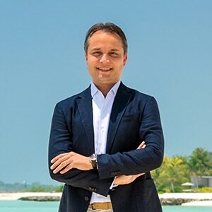 A portrait of Renato De Oliveira. He is a brown man with a dark blue suit standing at a beach.