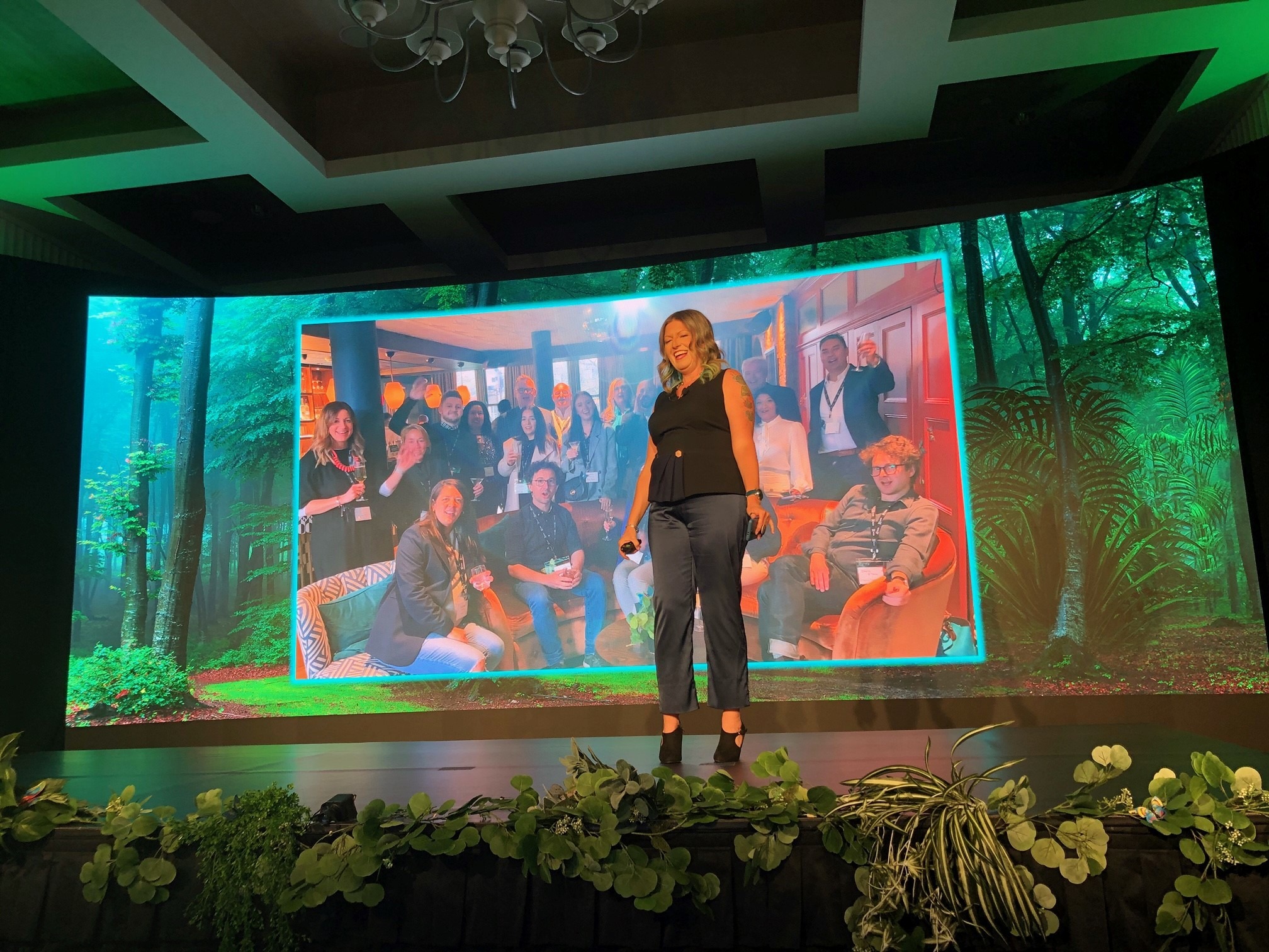 Rachel Moore stands, a blonde woman in black formal wear, on a stage decorated with faux plants in front of a projector