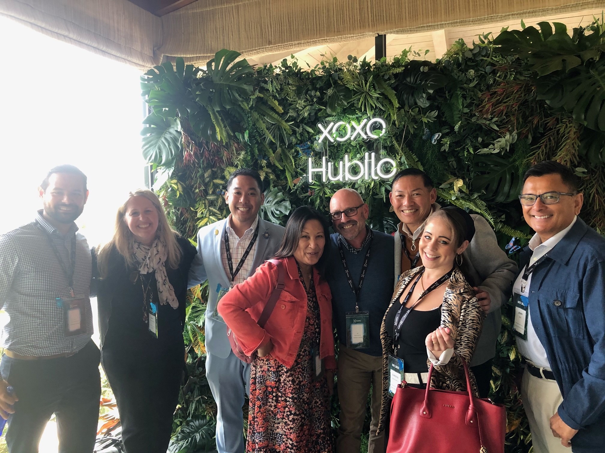 A group of attendees from Hubilo's MIX event standing in front of a Hubilo neon sign and faux plant wall.