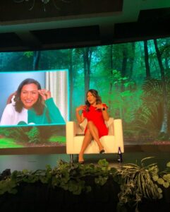 A woman in a red dress sits in a white armchair with another woman projected onto the screen behind her