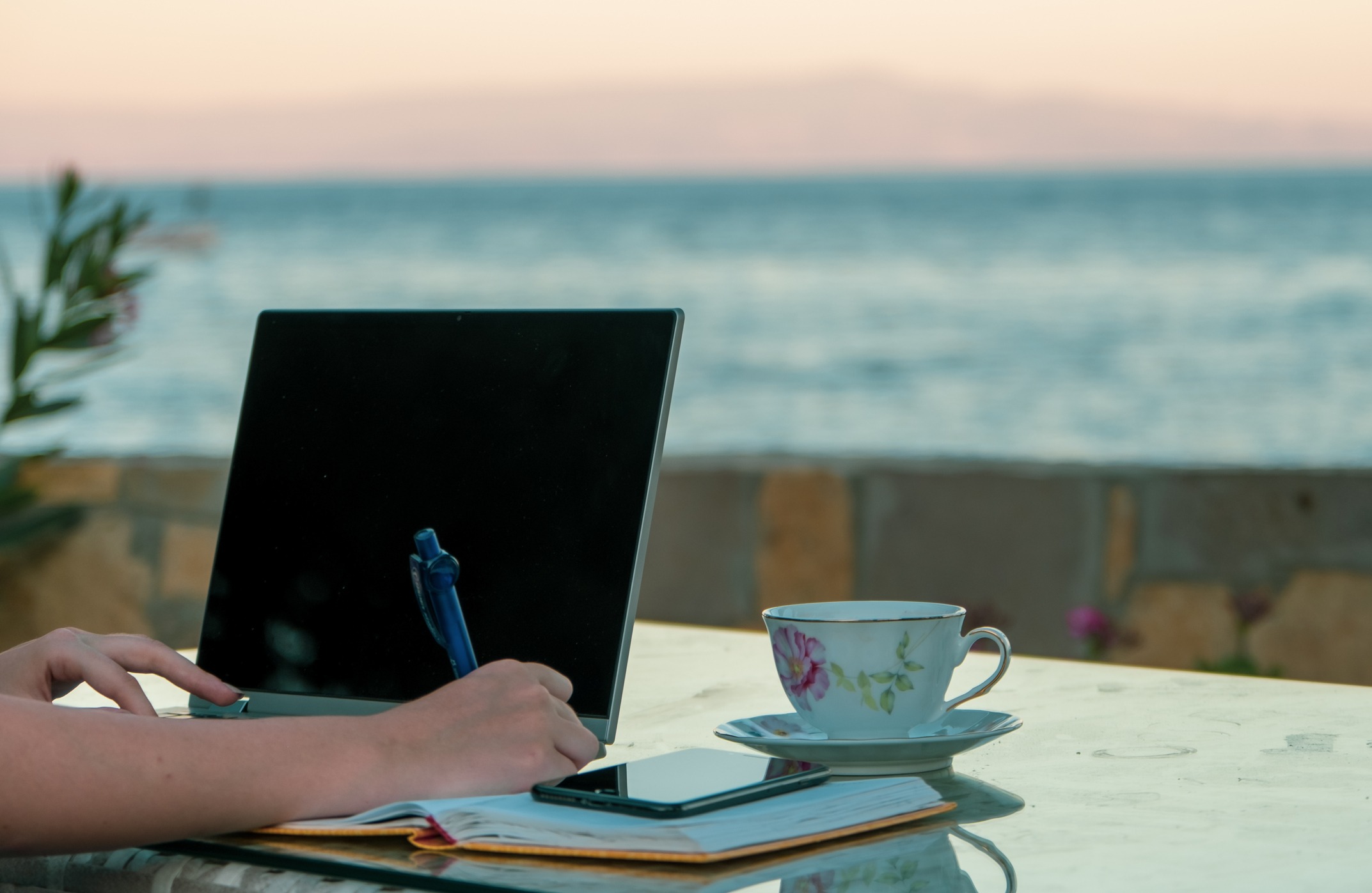 A person writing in a notebook with a laptop, phone and teacup on the table. A lake is in the background.