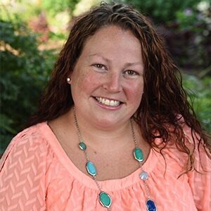 A portrait of Becca Lloyd. She is a white woman with wavy brown hair, a pink blouse and a turquoise necklace.