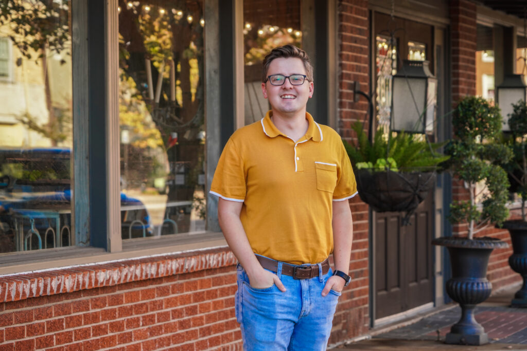 A portrait of Chris Riggins. He is a young white man with a yellow collared shirt in front of a brick storefront.