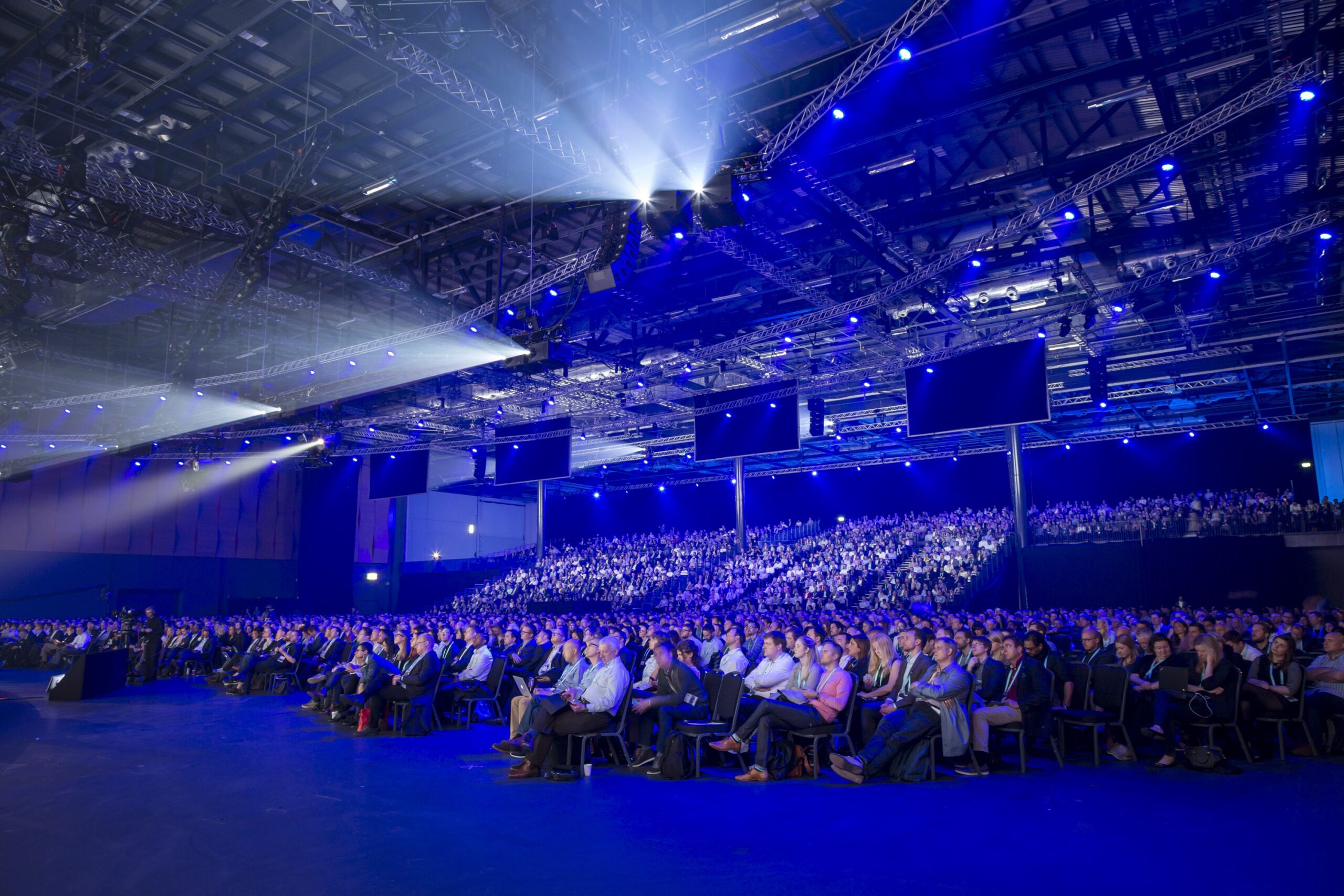 A large crowd in the ExCel event center. Stage lights above them give everything a blue tint.