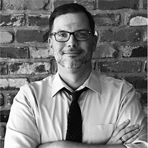 A black and white portrait of Jeff Carlson. He is a white man with square glasses, a white collared shirt and a dark tie.