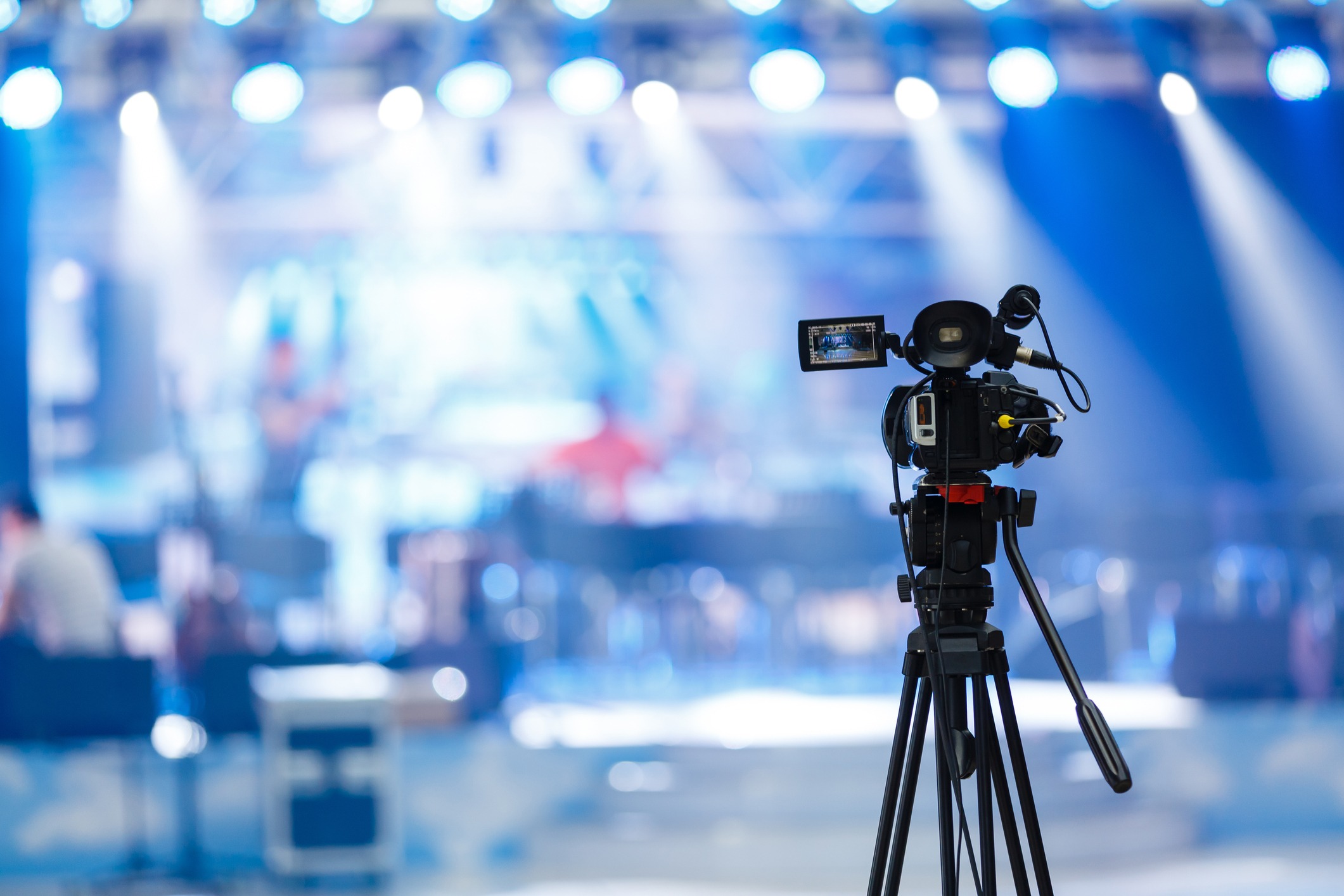 A TV camera set up on a tripod records a blurry stage in the background.
