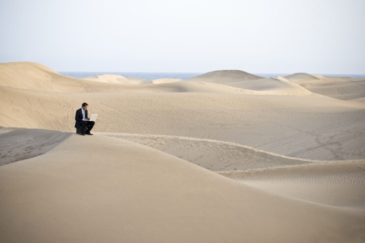 A businessman working on a laptop in the desert, representing remote work.