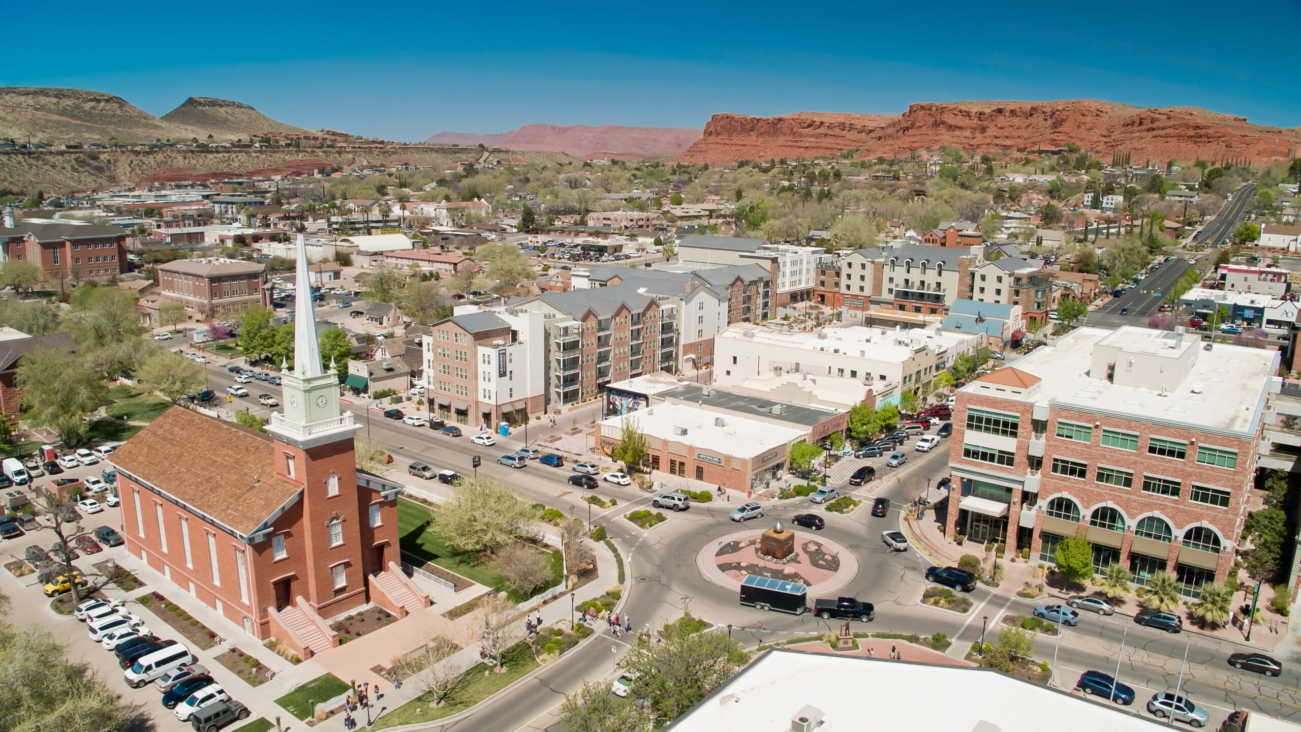 An aerial shot of St. George, Utah. Buildings are red brick with trees in between. A red bluff is in the distance.