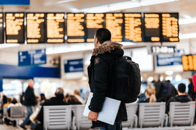 A man holding a laptop looks at a departure schedule. Practicing cybersecurity while traveling is important for all devices.