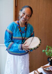 A portrait of Mayan healer Fernanda Montiel holding a healing instrument. She is wearing a blue striped blouse and a white lace skirt.