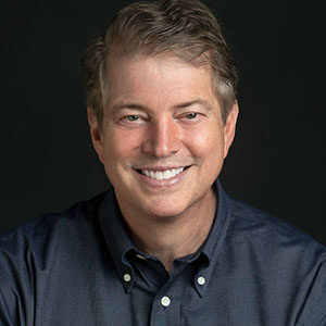 A portrait of Chris Caldwell. He is a white man with light combed hair and a dark collared shirt