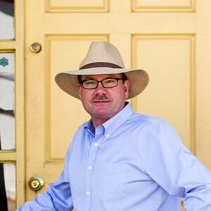 A portrait of Graeme Hughes. He is a white man with square glasses, a cowboy hat and a blue collared shirt.