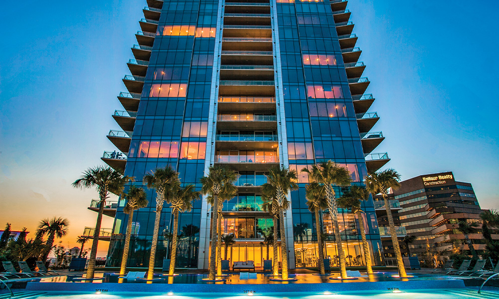 The exterior of the Harwood Boutique Hotel, a skyscraper with balconies on every floor and palm trees in front.