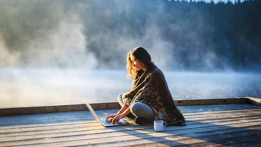 A woman works on her laptop while sitting next to a steaming lake.