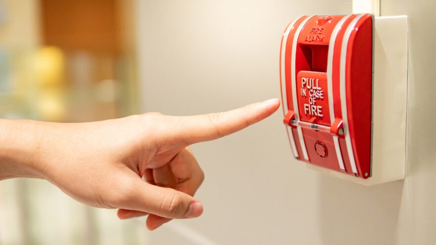 Male hand reaching for red fire alarm switch on concrete wall in office building