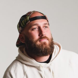 headshot of Cory Hymel Gigster, bearded and wearing a baseball cap and white sweatshirt