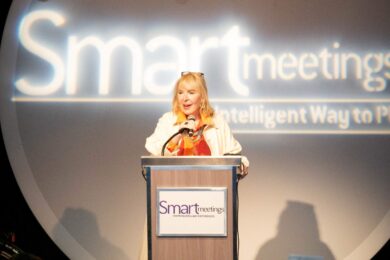 woman, marin bright, standing behind podium on stage in front of white neon "smart meetings" sign