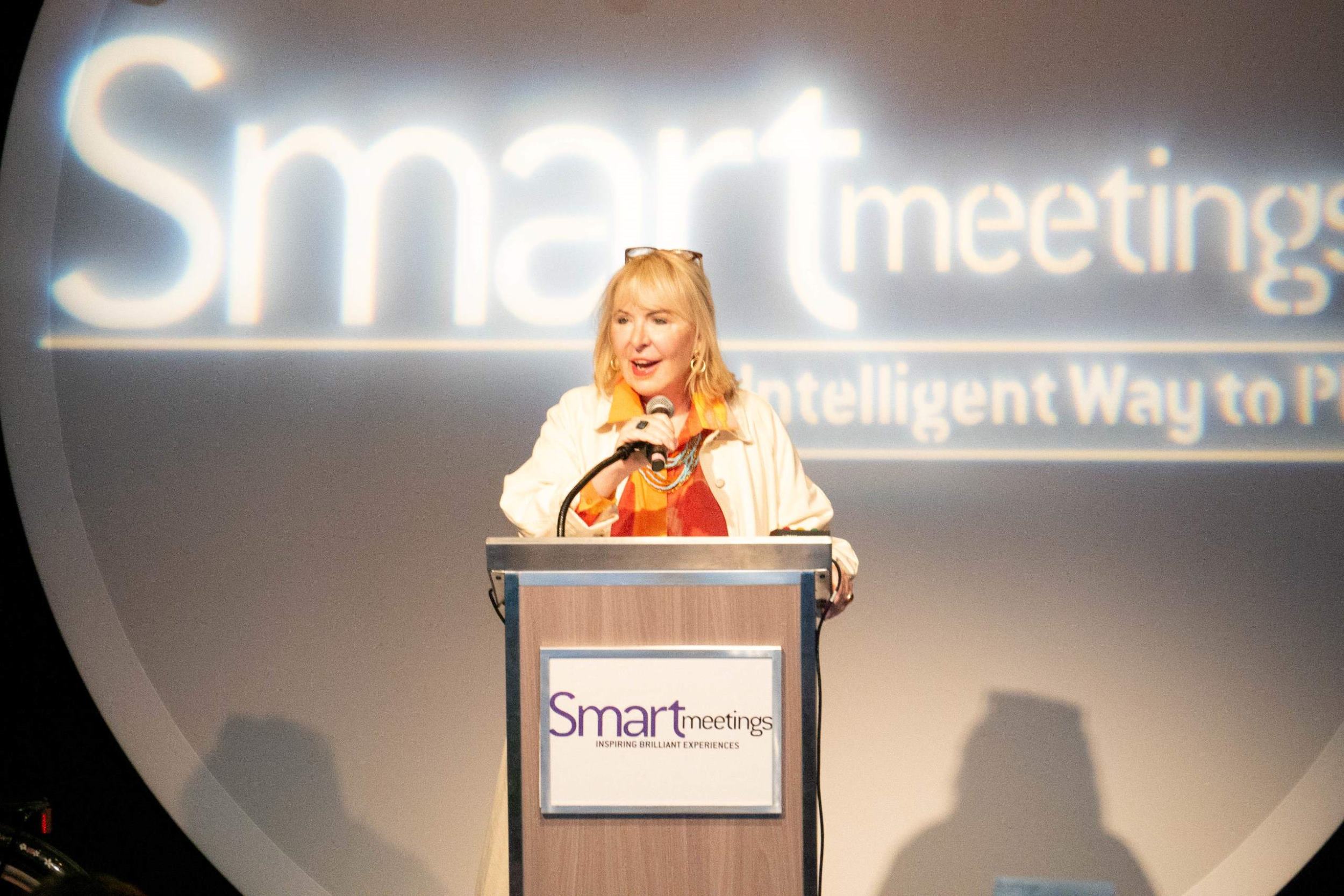 woman, marin bright, standing behind podium on stage in front of white neon "smart meetings" sign