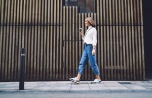 woman in jeans and sneakers strolling along street and drinking coffee